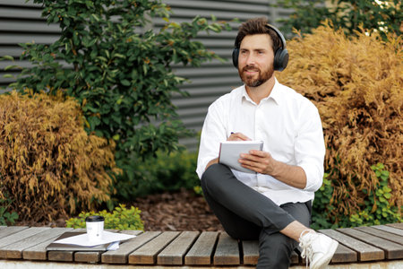 Handsome freelancer in headphones sitting on bench outdoors and writing business plan in notebook. Caucasian male thoughtfully looking aside while working in quiet place.の写真素材