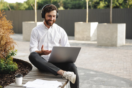Caucasian freelancer in wireless headphones working on portable laptop while sitting in urban area. Happy man discussing about new project and gesturing during online meeting.の写真素材
