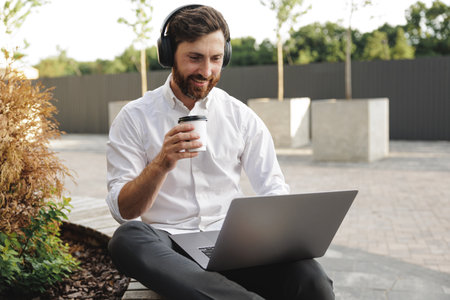 Front view of handsome businessman in headphones sitting on wooden bench and working outdoors. Smiling male holding coffee cup in hand while using portable laptop.の写真素材