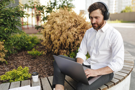 Handsome bearded businessman in formal clothing typing on portable laptop. Positive male in headphones attentively looking on screen while sitting on bench in urban area.の写真素材