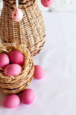 Easter composition with decorated tree branches in a wicker vase and pink colored eggs in wicker basket on white background. Copy spaceの写真素材
