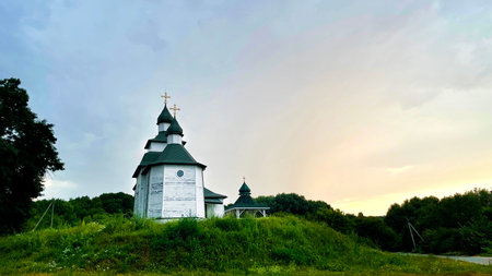 A wooden Orthodox church in the Cossack Baroque style, situated on a grassy hill in the village of Hryhorivka, Cherkasy region, Ukraine, during sunset.の写真素材
