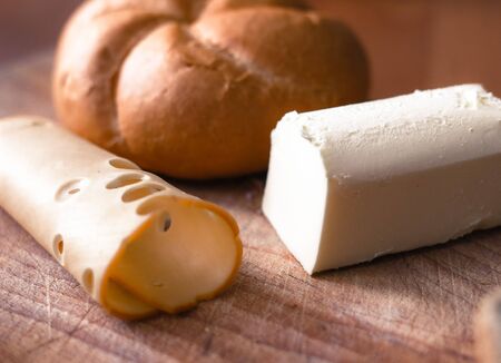 Freshly baked brown bread bunn, slice of smoked yellow cheese and piece of butter on wooden background. Healthy and tasty traditional breakfast or snackの写真素材