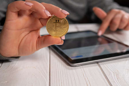 Closeup of a woman holding a golden bitcoin and working with charts on a tablet.