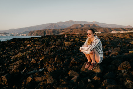 Charming girl in the sunglasses sits on the stone and waits for the sunset in the evening. Tenerifeの写真素材
