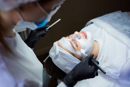 A cosmetologist side view who gently applies peeling gommage on the girlâs face. A doctor wearing medical face mask and disposable medical gloves during a procedure in a beauty salon for skin cleaningの写真素材