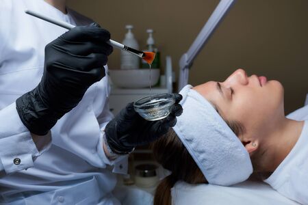 A female cosmetologist wearing black disposable medical gloves prepares to use a brush to apply a transparent face peeling to a young woman. A cosmetology procedure in a beauty salon for skin cleaningの写真素材