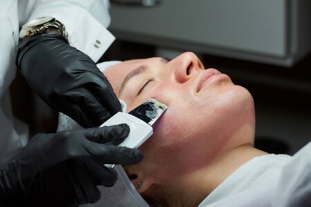 A close-up photo of the woman's face after a warming lotion during an ultrasonic procedure in the cosmetology office. A cosmetologist in disposable gloves peeling face with an ultrasonic skin scrubberの写真素材