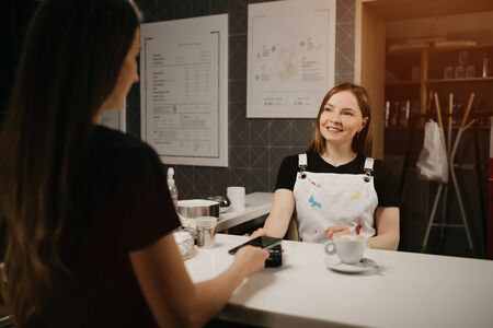 A female barista smiling holds out to a client a terminal for paying for a cup of coffee. A woman with long hair paying for a latte with a smartphone by contactless NFC technology in a cafe.の写真素材