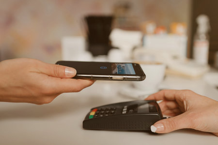 A close-up photo of a smartphone preparing to pay for a latte by contactless PAY PASS technology in a cafe. A female barista holds out a terminal for paying to a client in a coffee shop.の写真素材