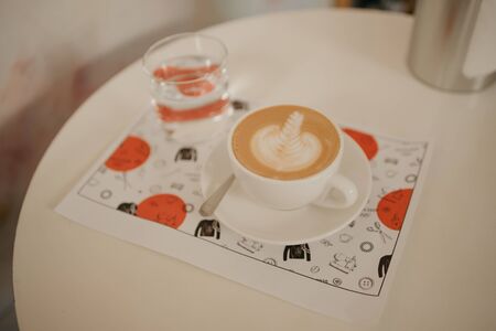 A cup of latte and a glass of water on a paper sheet on the white desk in a coffee shop. の写真素材