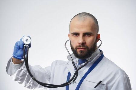 A serious doctor holding a stethoscope in his hand in a blue medical glove. A physician practitioner with a beard in a white lab coat and a blue tie.の写真素材