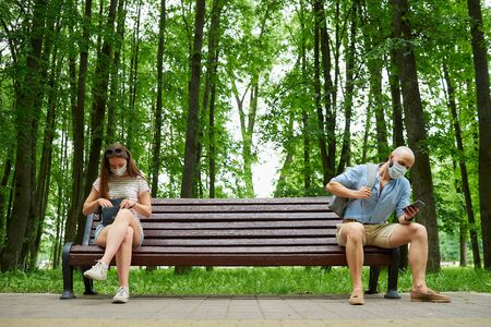 A bald man and a young woman sitting on opposite ends of the bench keeping distance one from another to avoid the spread of coronavirus. A guy and a girl in face masks in social distancing in the parkの写真素材