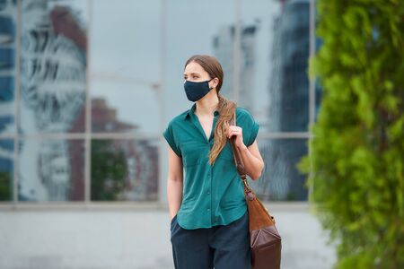 Cute woman in medical face mask walks thrusting hand into a pocket of trousers, holds a bag in downtown. Girl keeping social distance wears a protective face mask to avoid the spread of coronavirusの写真素材