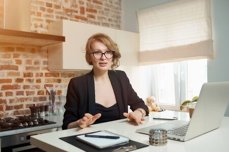 A young woman in glasses works remotely on a laptop in her kitchen. A blond girl gesticulating discusses with her colleagues on a video conference at home. Lady teaching a lecture online on a webinarの写真素材