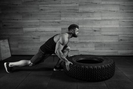 A bodybuilder with a tattoo and beard is pushing a tire in a gym. A muscular shredded man in a black tank top and space gray shorts is doing a crossfit workout. A black and white photo.の写真素材