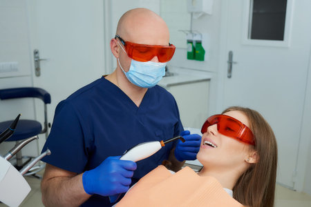 A dentist in a medical face mask and UV protection glasses holding a dental polymerization light...