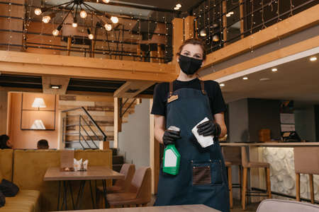 A waitress who wears a black medical face mask and disposable medical gloves is holding a bottle with sanitizer and a rag to clean tables in a restaurant. A barista is waiting for clients in a cafe.の写真素材