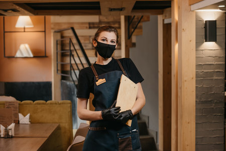 A waitress in black disposable medical gloves wears a medical face mask is holding a wooden menu in...