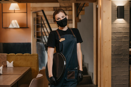 A waitress in black disposable medical gloves wears a medical face mask is holding a plastic tray in...