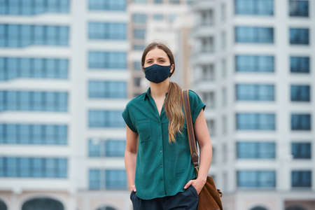 A portrait of a woman in a medical face mask is thrusting hands into pockets of trousers while walking in the center of the city. A girl in a protective mask keeping social distance on the streetの写真素材