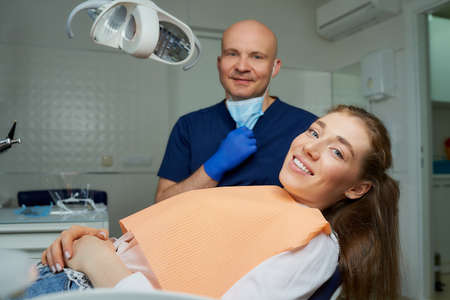 A bald dentist lowered the medical face mask near a laying female patient in dental chair. A doctor with a patient who is demonstrating an excellent result of the tooth treatment in a dentist's officeの写真素材