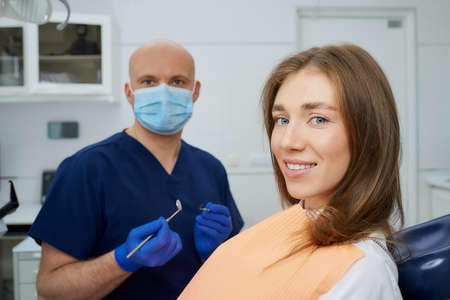 A bald dentist in a medical face mask is holding a dental mirror and dental explorer near a cute patient in a dental chair. A doctor with a patient who is demonstrating a result of the tooth treatmentの写真素材