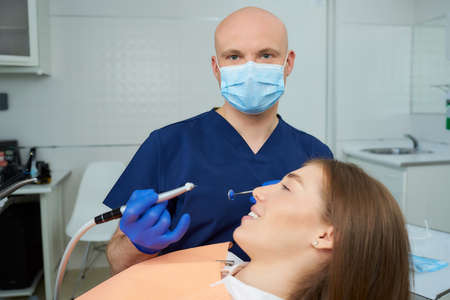 A bald dentist in a medical face mask holds a dental mirror and drill near a lying young woman in the dental chair. A doctor and his female patient during a dental procedure in a dentist's office.の写真素材