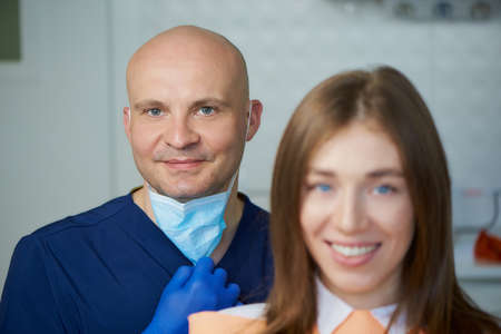 A bald smiling middle-aged dentist together with his young female patient in a dentist's office. A doctor and his patient are demonstrating an excellent result of the teeth treatment.の写真素材