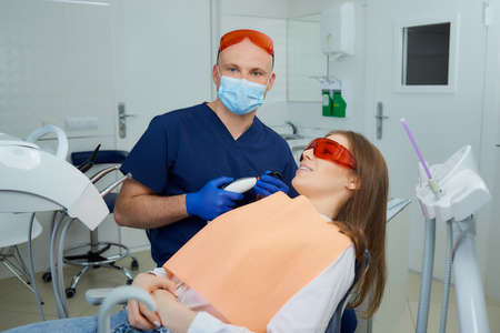 A dentist in a medical face mask and UV protection glasses holding a dental polymerization light near a girl in the dental chair. A doctor and a female patient after treatment in a dentist's office.の写真素材