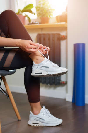 A close photo of the hands and legs of a sporty girl in black tight suit who is tying her shoelaces on white sneakers while sitting on the chair before training at home.の写真素材