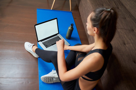 A sideways photo of a sporty girl in black tight pants and top is sitting on the blue yoga mat with...