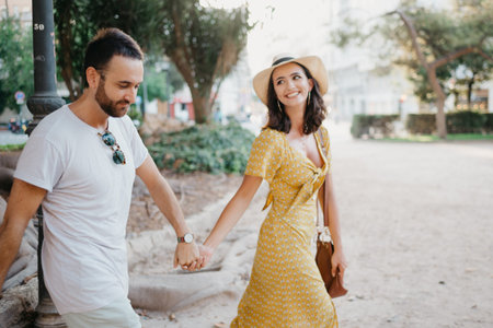 A smiling girl in a hat and a yellow dress and her boyfriend with a beard are walking holding each other's hand in a park in Spain. A happy couple of lovers are enjoying Valencia in the evening.の写真素材