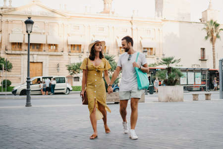 Girl in a hat and a dress with a plunging neckline and her boyfriend with a beard and sunglasses are relaxing holding each other's hand in the old town. A couple of tourists on the sunset in Valencia.の写真素材