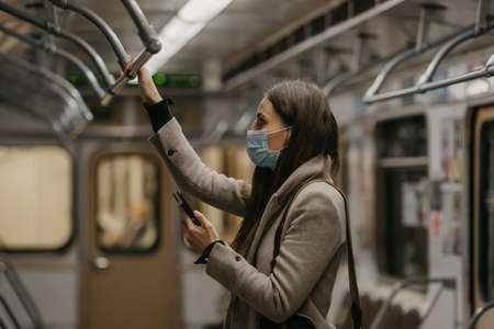 A woman in a medical face mask to avoid the spread of coronavirus is dreaming in a subway car. A girl in a surgical mask on her face against COVID-19 is holding a cellphone on a train.の写真素材