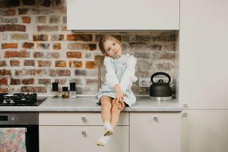 A happy young blonde girl with long hair is posing while sitting on the table in the kitchen.の写真素材