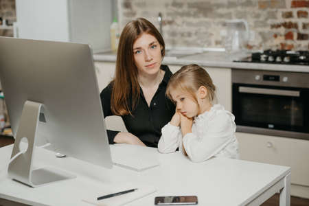 A mother is posing at her workplace near a computer while her daughter is bored at home. A businesswoman is working remotely from her apartment.の写真素材