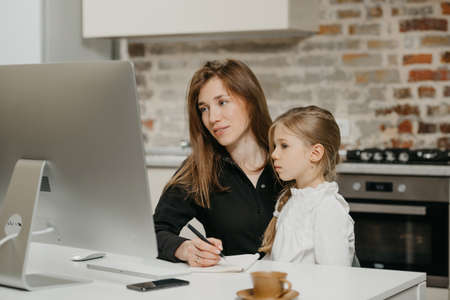 A young mom is working remotely at home while a daughter is staring at a computer. A pretty businesswoman is taking notes while working from an apartment near her blonde child.の写真素材