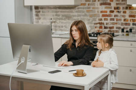 A mother is working remotely at home while a daughter is watching it. A businesswoman is working from an apartment near her blonde child.の写真素材