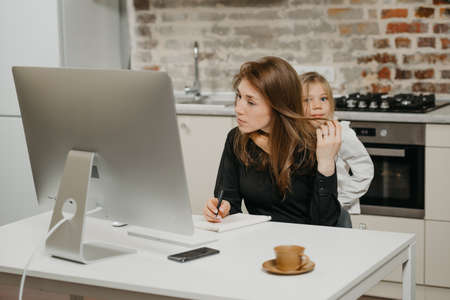 A young mother is working remotely at home while a daughter is hugging her. A businesswoman is doing notes while working from an apartment near her blonde child.の写真素材