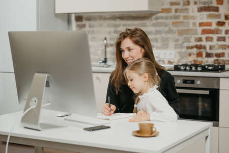 A young mother is working remotely at home while a daughter is studying from her. A smiling businesswoman is taking notes while working from an apartment near her blonde child.の写真素材