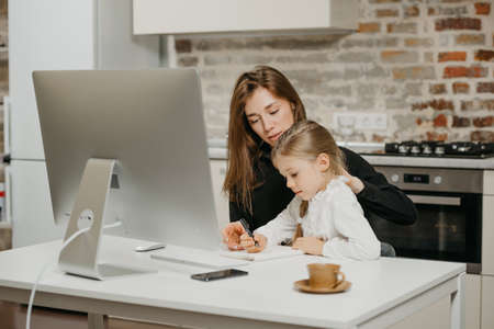 A young mother is teaching a daughter at her remote workplace at home. A pretty businesswoman is describing to her blonde child some things.の写真素材