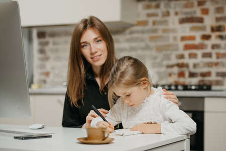A mom is posing and hugging a pretty daughter while she is drawing in the notebook at the workplace. A gorgeous mother watches her blonde child which is doing homework at home.の写真素材