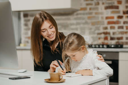 A young smiling mom is hugging a pretty daughter while she is writing in the notebook at the workplace. A gorgeous mother is looking at her blonde child which is doing homework at home.の写真素材