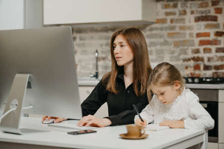A young mom is working remotely carefully while a daughter is drawing in the notebook at home. A pretty mother is reading news on a computer near her blonde child which is writing diligently.の写真素材