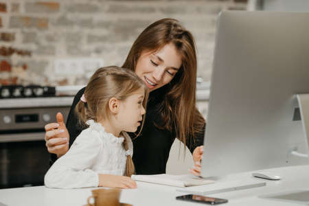 A young smiling mother is teaching a daughter at her remote workplace at home. A pretty mom is describing to her blonde child some things.の写真素材