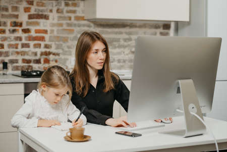 A young mom is working remotely carefully while a daughter is drawing in the notebook at home. A pretty mother is studying on a computer near her blonde child which is writing letters diligently.の写真素材
