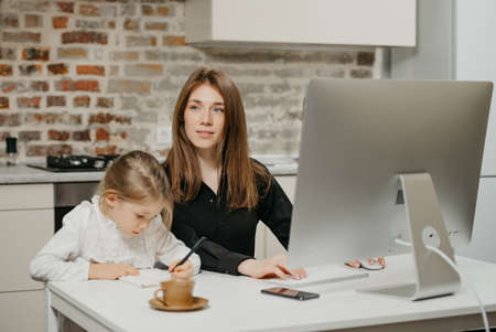 A young mom is staring away while a daughter is drawing in the notebook at home. A pretty mother is working remotely on a computer near her child which is writing letters diligently.の写真素材