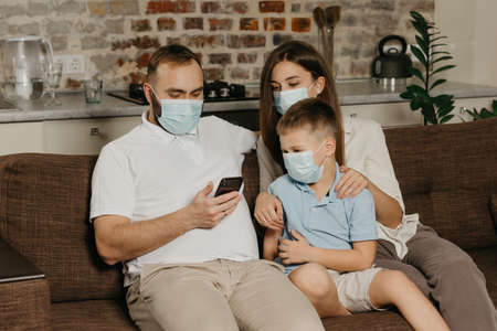 Father, mother and son are sitting on a sofa in medical face masks to avoid the spread of coronavirus (COVID-19). Family during quarantine at home. Dad is watching news on a phone near his relativesの写真素材