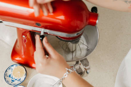 The hands of a woman who is switching speeds of a red stand mixer which is whipping the egg whites in a stainless steel soup bowl in a kitchen.の写真素材